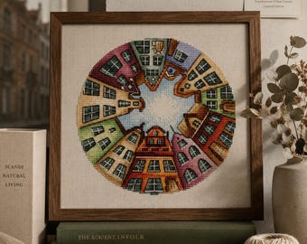Puede incluir: Un marco de madera cuadrado que contiene un bordado de punto de cruz circular con casas de colores dispuestas en círculo. Los edificios presentan diversos estilos arquitectónicos y colores, como rojo, amarillo, verde y azul, rodeando un cielo blanco.