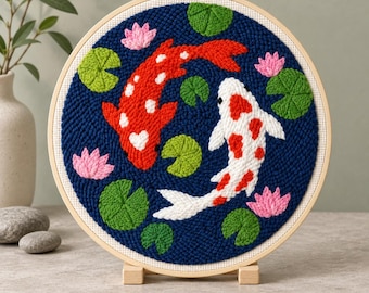 Puede incluir: Un bastidor de bordado circular con un diseño de aguja mágica que muestra un pez koi rojo y un pez koi blanco con manchas rojas nadando en un estanque azul oscuro, rodeados de nenúfares verdes y pequeñas flores de loto rosas.