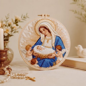 May include: A circular embroidered artwork in a wooden hoop. It shows a woman in a blue robe holding a baby in a woven basket, surrounded by cherubs. The background has a golden halo design, with a soft, neutral colour palette.