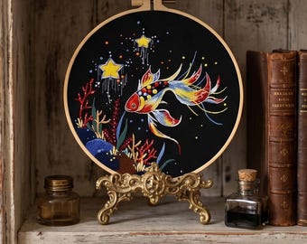 May include: A circular embroidery hoop featuring a black fabric background with a colorful fish swimming among coral and yellow stars. The fish has red, yellow, and blue fins. The embroidery is displayed on an ornate gold metal stand.