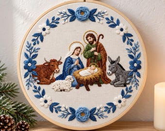 Puede incluir: Un bastidor de bordado circular que muestra una escena de natividad con María, José y el niño Jesús en un pesebre. La escena incluye un buey marrón, un burro gris y un cordero blanco, rodeados por una corona de flores azules y blancas.