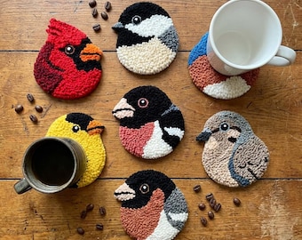Puede incluir: Una colección de seis posavasos circulares de punch needle con forma de varios pájaros, incluyendo un cardenal rojo, un pinzón amarillo y otros con plumaje negro, blanco, gris y rosa, dispuestos con dos tazas de cerámica y granos de café.