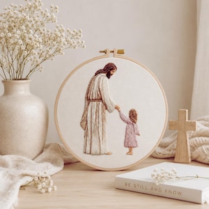 Pode incluir: Um bastidor de bordado circular com uma ilustração costurada de uma figura com uma túnica longa segurando a mão de uma criança pequena com um vestido rosa. O tecido é branco sujo e o bastidor é de madeira clara com um parafuso de metal.