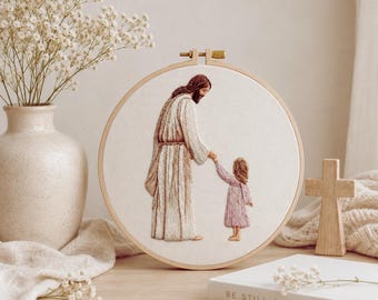 Puede incluir: Un bastidor de bordado circular que presenta una ilustración cosida de una figura con una túnica larga que sostiene la mano de un niño pequeño con un vestido rosa. La tela es de color blanco roto y el bastidor es de madera clara con un tornillo.