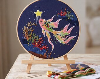 May include: A circular embroidery hoop containing a dark blue fabric with a hand-stitched design of a multi-colored fish swimming toward a yellow star. The scene includes embroidered coral and seaweed. A wooden easel holds the hoop, with embroidery floss.