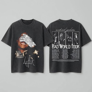 May include: A charcoal gray short-sleeve t-shirt featuring a graphic of Michael Jackson wearing a sequined glove on the front, with a signature below. The back displays four black and white photos, the text "michael jackson BAD WORLD TOUR", and a list of tour dates.