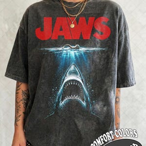 Op de afbeelding: Een antracietgrijs acid-wash T-shirt met het rode JAWS-filmlogo boven een afbeelding van een zwemmer aan het wateroppervlak en een grote haai met een open bek die uit het diepblauwe water omhoog komt. Het T-shirt heeft korte mouwen.