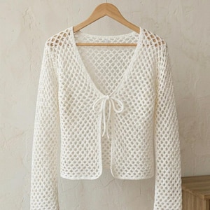 Op de afbeelding: Een witte, open geweven gehaakte cardigan met lange mouwen en een V-hals. De cardigan heeft een striksluiting bij de borst en hangt aan een houten hanger. Het haakpatroon creëert een delicate, luchtige textuur.