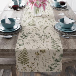 May include: A beige table runner featuring a botanical print of green ferns, eucalyptus leaves, and wildflowers. The runner is centered on a wooden table set with teal bowls, white napkins, wine glasses, and a vase of pink lilies.
