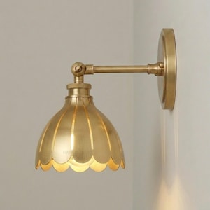 Peut inclure: Une applique murale en laiton dotée d'un abat-jour en forme de cloche avec un bord festonné. Le luminaire comprend une plaque murale circulaire, un bras horizontal avec une articulation pivotante et une finition métallique. HAYATSTUDIO est inscrit.