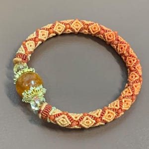 May include: A handcrafted bracelet featuring a woven design in shades of peach and tan, adorned with small coral-colored beads. The bracelet includes a large, round amber-colored stone accented with gold-colored filigree and clear crystals. The bracelet is a jewelry item.