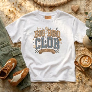 Könnte beinhalten: Weißes T-Shirt mit "BIG BRO CLUB"-Grafik in Braun und Blau, mit Sternen, Blitzen und Smiley-Gesichtern. Das Shirt enthält "EST. 2024" auf einem Banner. Braune und weiße Turnschuhe und grüne Hosen sind ebenfalls zu sehen.