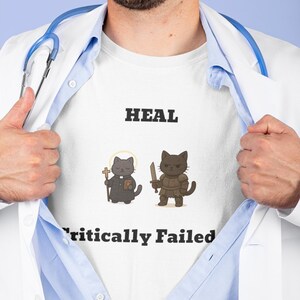 May include: White t-shirt with the word "HEAL" above two cartoon cats. One cat is dressed as a priest, the other as a warrior. The text "Critically Failed" is below the cats. The shirt is being revealed by a person.