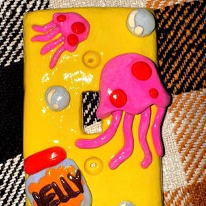 May include: A rectangular yellow light switch cover plate featuring three-dimensional clay decorations including two pink jellyfish with red spots, three light blue bubbles, and a small jar labeled JELLY with a red lid and orange contents.