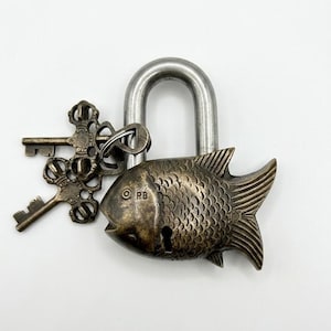 May include: A brass padlock shaped like a fish with a silver-toned metal shackle and two ornate keys attached to a ring. The fish body features detailed scales and the letters PB stamped near the eye. The keys have intricate, decorative heads.