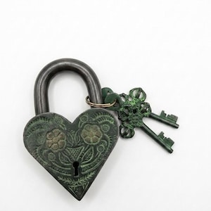 May include: A heart-shaped metal padlock with a dark, weathered green patina finish featuring embossed floral and leaf patterns on the front. A keyhole is centered at the bottom, and two ornate, matching keys are attached to the shackle.