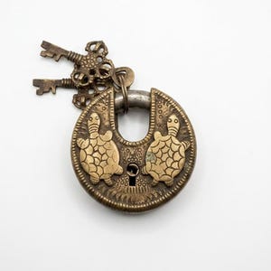 Può includere: Un lucchetto circolare in ottone con due figure di tartaruga in rilievo sul davanti e una serratura centrale. Due chiavi in ottone ornate e abbinate sono fissate all'arco superiore. Il metallo presenta una patina invecchiata e dettagliata.