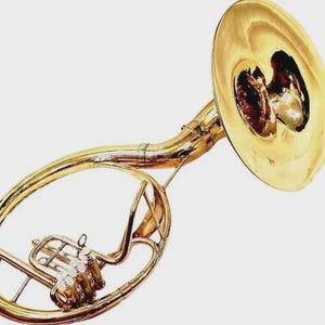 Op de afbeelding: Een gepolijste koperen mars-hoorn met een grote, brede beker, een gebogen buisstructuur en drie verticale ventielen met witte vingerknoppen. Het instrument heeft een heldere, reflecterende metalen afwerking over het gehele oppervlak.