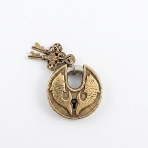 Peut inclure: Un cadenas circulaire en laiton orné de deux poissons en relief se faisant face avec une serrure centrale. Une anse en métal argenté relie le cadenas à un anneau décoratif en laiton portant deux petites clés finement travaillées.