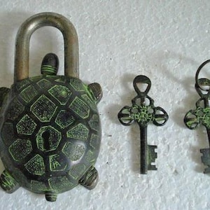 May include: A turtle-shaped padlock with a dark green and black finish, featuring a metallic shackle and a keyhole on the shell. Two matching ornate keys with intricate cutouts and a green patina finish are positioned to the right of the lock.