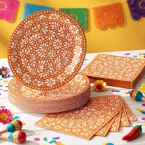 May include: A stack of round orange paper plates and a stack of square orange paper napkins, both featuring a repeating white floral and leaf pattern. The items are arranged on a white surface with scattered colorful confetti and small maracas.