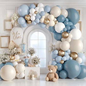 May include: A large balloon arch featuring a mix of matte blue, white, cream, and metallic gold balloons. A large white balloon on the left displays the cursive text 'Nlor Qia's'. A light brown teddy bear sits on the floor near the arch base.