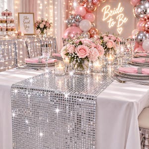 May include: A silver mirrored mosaic table runner made of small square tiles, draped over a white tablecloth. The runner features a reflective surface that catches light, creating a sparkling effect on a table set with pink floral centerpieces.