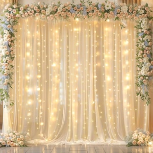May include: A rectangular backdrop featuring sheer cream-colored fabric curtains illuminated by vertical strings of warm white fairy lights. The frame is adorned with a dense floral arrangement of pale pink roses, white flowers, and blue accents.