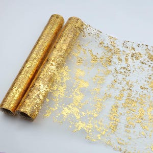 May include: A roll of sheer fabric featuring a metallic gold foil pattern. The material is translucent with an irregular, textured gold leaf design applied across the surface, creating a shimmering effect that contrasts with the light base.