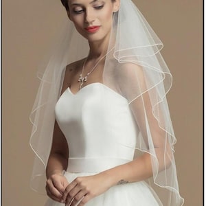 May include: A two-tier white bridal veil featuring a delicate rolled edge finish. The sheer tulle fabric drapes over the head and shoulders, complementing a strapless white gown and a silver-toned necklace with clear crystal accents.