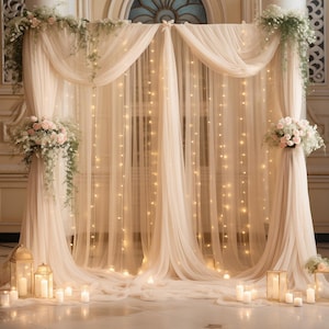 May include: A decorative backdrop featuring layers of sheer cream-colored fabric draped over a frame, illuminated by vertical strings of warm white fairy lights. The structure is adorned with clusters of white flowers and greenery at the top corners.