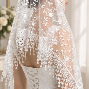 May include: A white lace veil featuring intricate floral embroidery and a scalloped edge with dangling circular accents. The veil is draped over the head and shoulders, showcasing a delicate mesh fabric with detailed needlework patterns throughout.