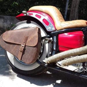 Peut inclure: Une moto personnalisée avec une sacoche de selle en cuir marron, une selle beige et un cadre rouge. La moto a un pneu à flancs blancs et des composants de moteur chromés. Les tuyaux d'échappement sont enveloppés dans un matériau beige.
