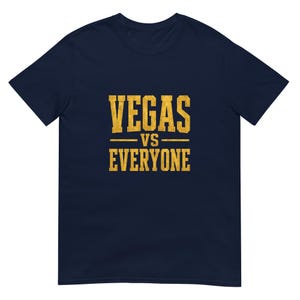 Op de afbeelding: Een marineblauw t-shirt met korte mouwen en de tekst "VEGAS VS EVERYONE" in een verweerd, goudgeel bloklettertype in het midden op de borst. Het shirt heeft een ronde hals en een standaard, recht model.