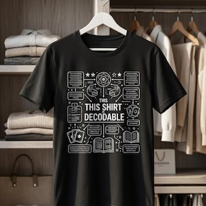 Op de afbeelding: Een zwart t-shirt met korte mouwen en een witte grafische print met de centrale tekst "THIS SHIRT IS DECODABLE", omringd door diverse lijntekeningen zoals een atoom, speelkaarten en open boeken met bijbehorende onleesbare tekstblokken.