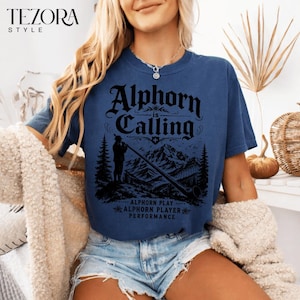 May include: A navy blue t-shirt with the text "Alphorn is Calling" in a black, stylized font. The graphic features a silhouette of a person playing an alphorn in a mountain landscape. The shirt also has the text "Alphorn Play, Alphorn Player, Performance".