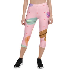 Puede incluir: Mallas capri rosas con un estampado de macarons coloridos y pequeños lunares rosas. Los macarons son en tonos morado, verde, naranja y marrón. Las mallas se llevan con zapatillas deportivas negras.