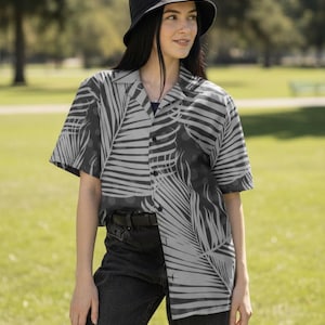 Peut inclure: Une personne porte un chapeau de pêcheur noir et une chemise boutonnée à manches courtes. La chemise présente un motif de feuilles de palmier gris et noir. La personne porte également un jean noir.