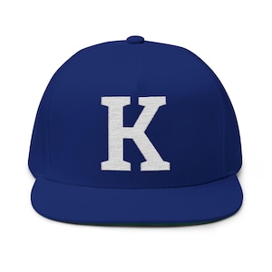 May include: A royal blue flat-brimmed baseball cap with a white, block letter "K" embroidered on the front. The cap has a structured crown, a flat brim, and a green underside. A simple, stylish accessory.