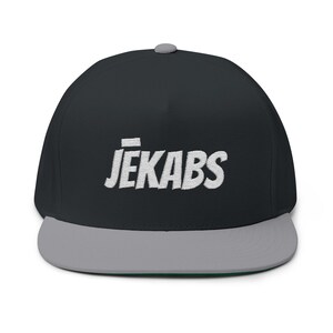 Può includere: Cappellino da baseball nero a tesa piatta, con visiera grigia e bottone grigio. Il testo bianco ricamato "JEKABS" è sulla parte anteriore del cappellino.