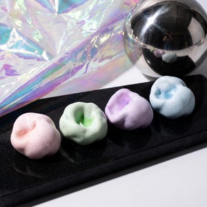 May include: Four small, rounded, matte-textured objects in pastel colors: light pink, pale green, soft lavender, and baby blue. Each piece features a central indentation, resembling a stylized tooth or organic form, arranged in a row.