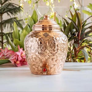 May include: A copper water dispenser featuring an intricate embossed floral pattern across its rounded body. The vessel includes a matching lid with a knob handle and a small spigot at the base with a red circular accent. The surface is polished.