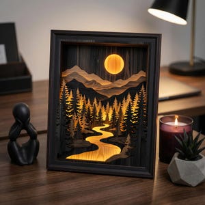 May include: A rectangular wooden shadow box art piece featuring a layered landscape of dark silhouettes of pine trees and mountains against a glowing yellow moon and a winding river. The internal light illuminates the scene from behind the cutouts.
