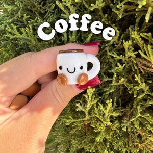 May include: A small white ceramic mug charm with a brown rim and base, featuring a hand-painted black smiling face with two eyes and a curved mouth. The word coffee is written in white letters above the mug.