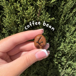 May include: A small, smooth, brown coffee bean charm with a glossy finish, featuring a hand-painted smiling face with two black eyes and a small mouth, along with the text coffee bean written in white letters above the object.