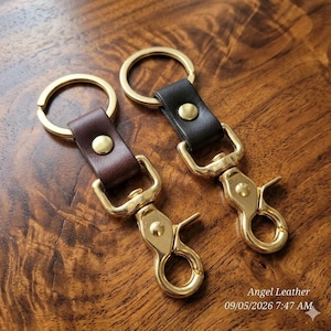 May include: Two keychains featuring a gold-toned metal split ring and a lobster clasp connected by a leather strap. One strap is dark brown and the other is black. Each strap is secured with a gold-toned metal rivet. Text reads Angel Leather.