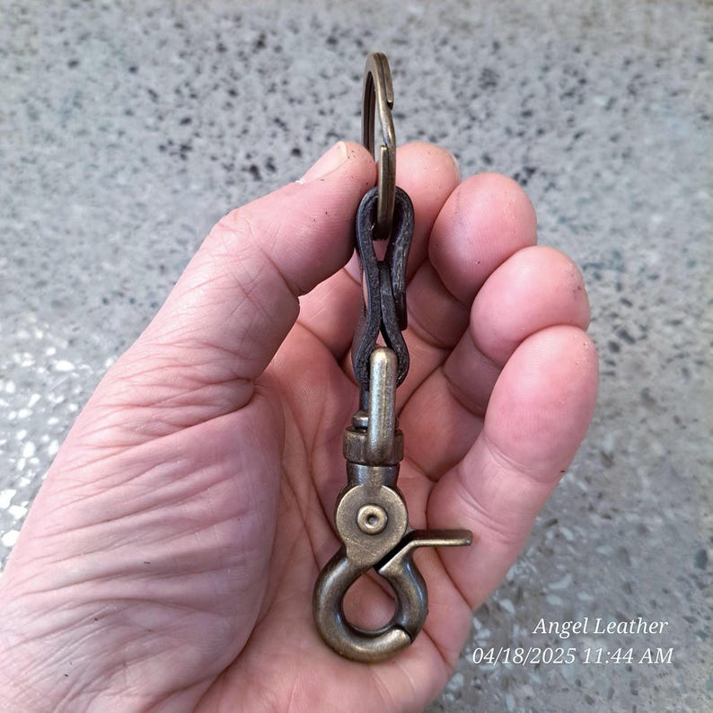 May include: A brass key chain with a leather loop and a spring clasp. The key chain is held in a hand.