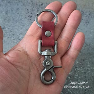 May include: A keychain featuring a dark red leather strap secured with a silver-toned metal rivet, attached to a circular split ring at the top and a heavy-duty, swivel-style lobster claw clasp in a matching brushed silver-toned metal finish.