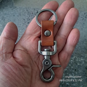 May include: A tan leather keychain featuring a circular metal ring attached to a rectangular leather strap with a silver-toned snap button. The strap connects to a swivel lobster clasp made of brushed dark silver metal hardware.