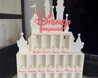 Puede incluir: Un expositor blanco de varios niveles con forma de castillo, con el texto rojo Disney Playmobil en la parte superior. El soporte tiene múltiples compartimentos rectangulares para figuras, con nombres como Aladin, Genio, Gepeto y Pinocho en rojo.